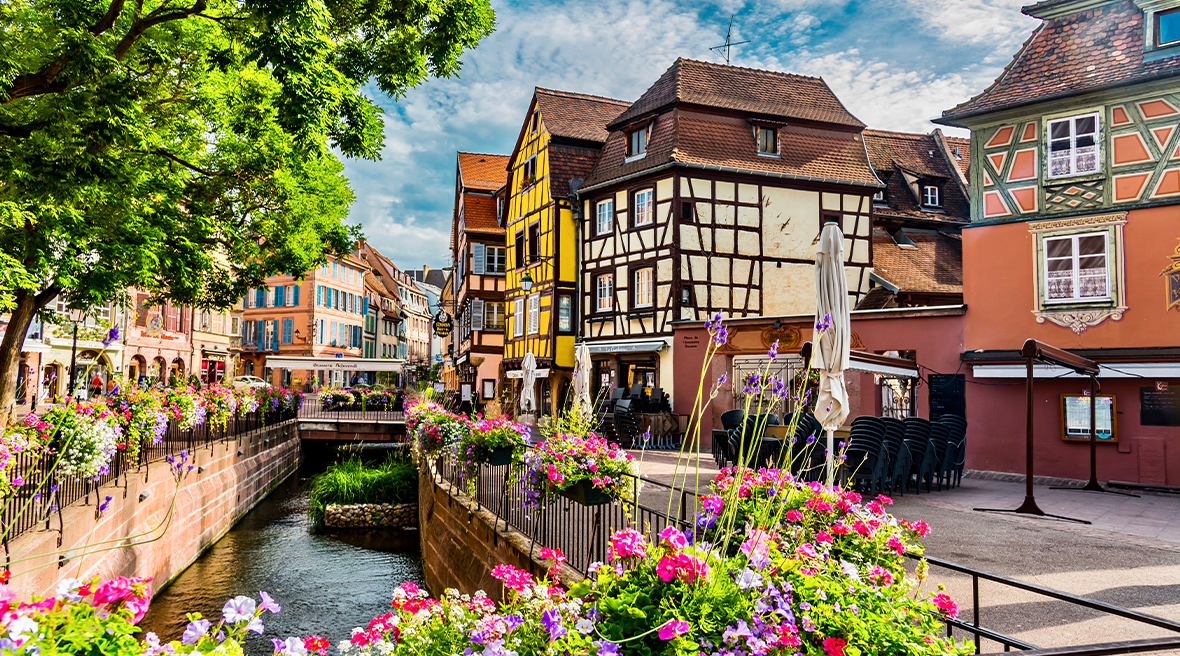brightly coloured timbered houses lining a river with paths each side and flowers lining a bridge across the water